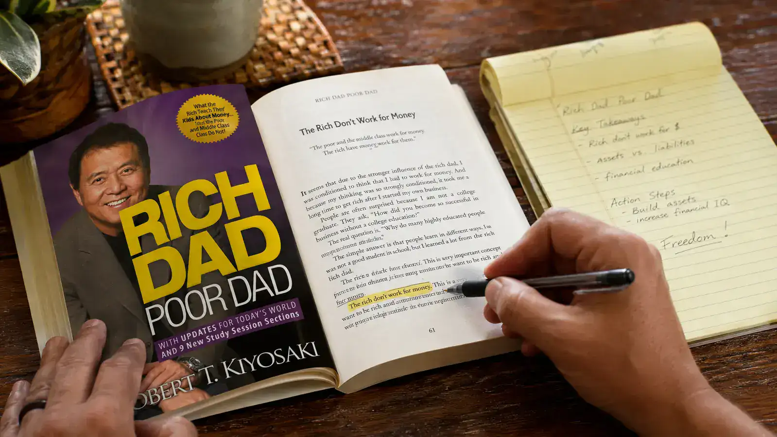 Close-up of the Rich Dad Poor Dad book cover resting on a wooden table next to a pen and a notepad with handwritten notes